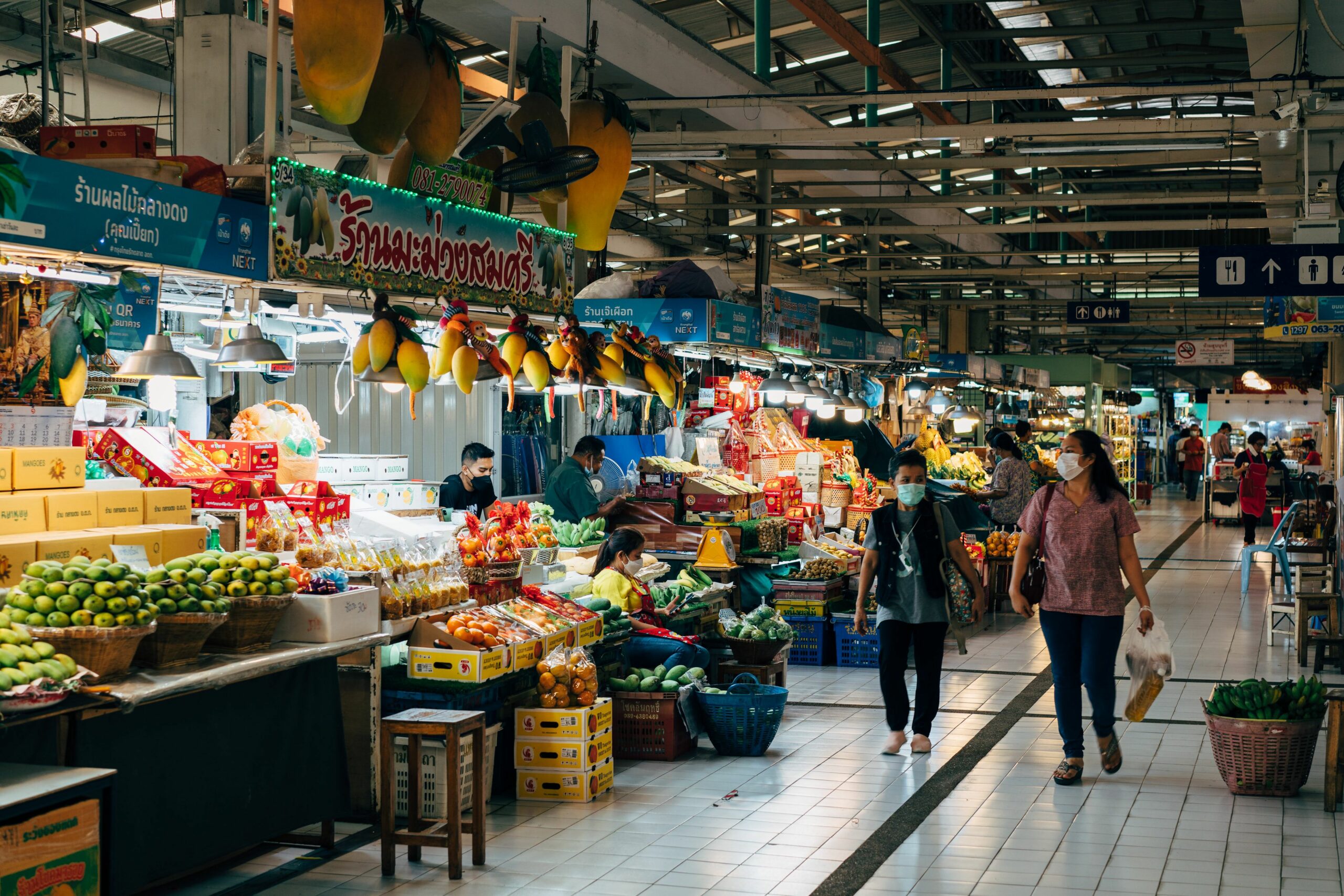 Asian indoor market with fruit stalls and shoppers wearing masks, showcasing vibrant produce.