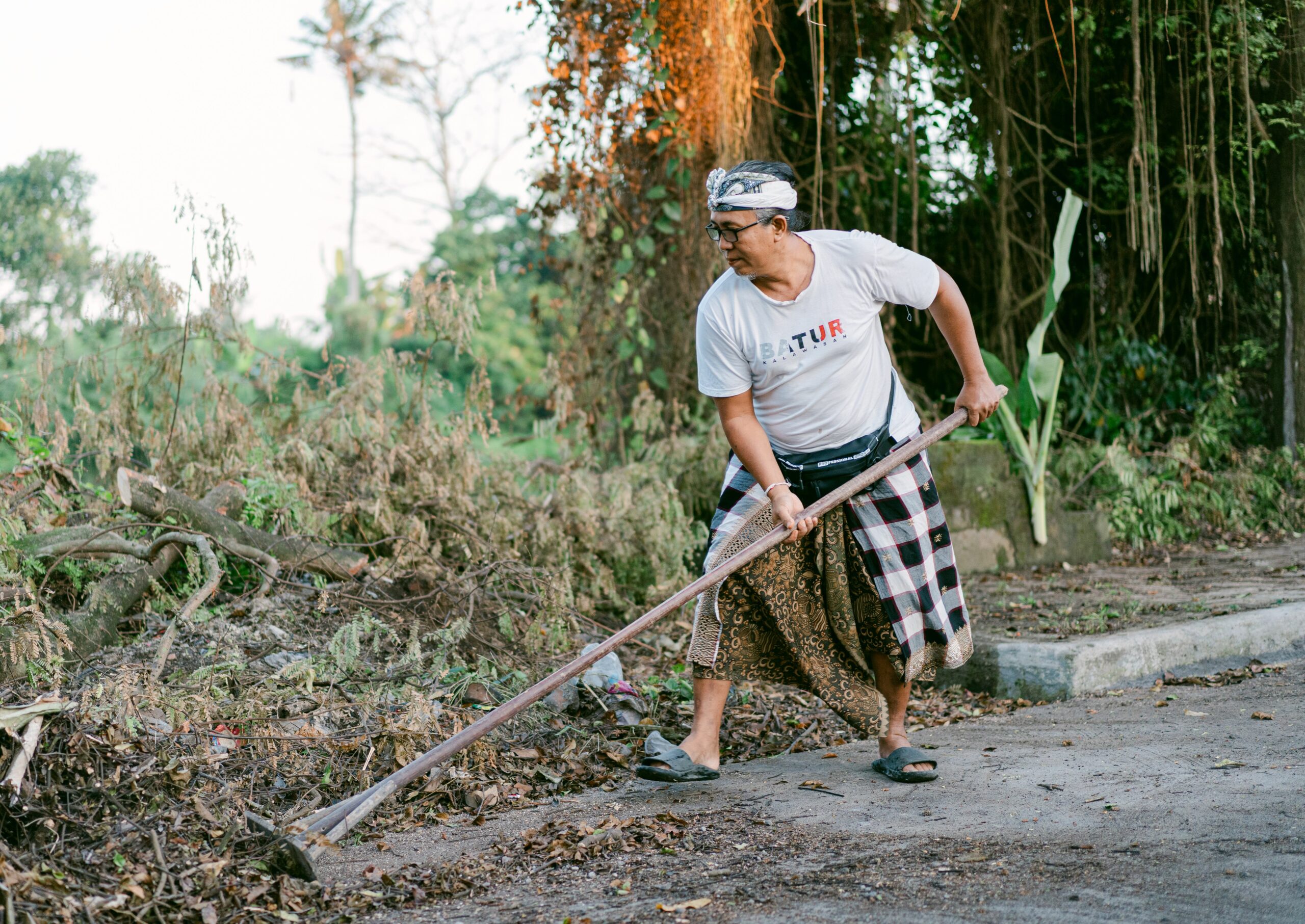 A Balinese man rakes leaves outdoors while wearing traditional attire, embodying cultural practices.