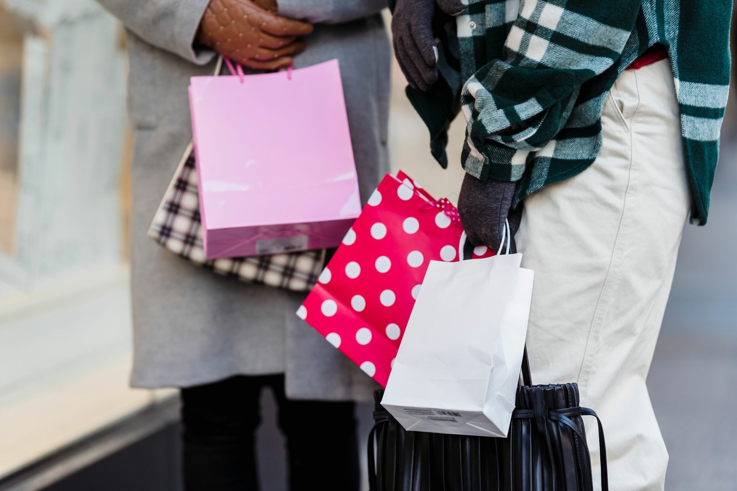 Unrecognizable female friends with various colorful shopping bags standing on sidewalk near showcase on street in city on blurred background